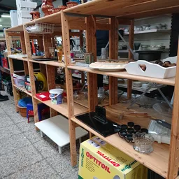 Houten stellingen met diverse spullen en voorwerpen in een kringloopwinkel, met een persoon op de achtergrond