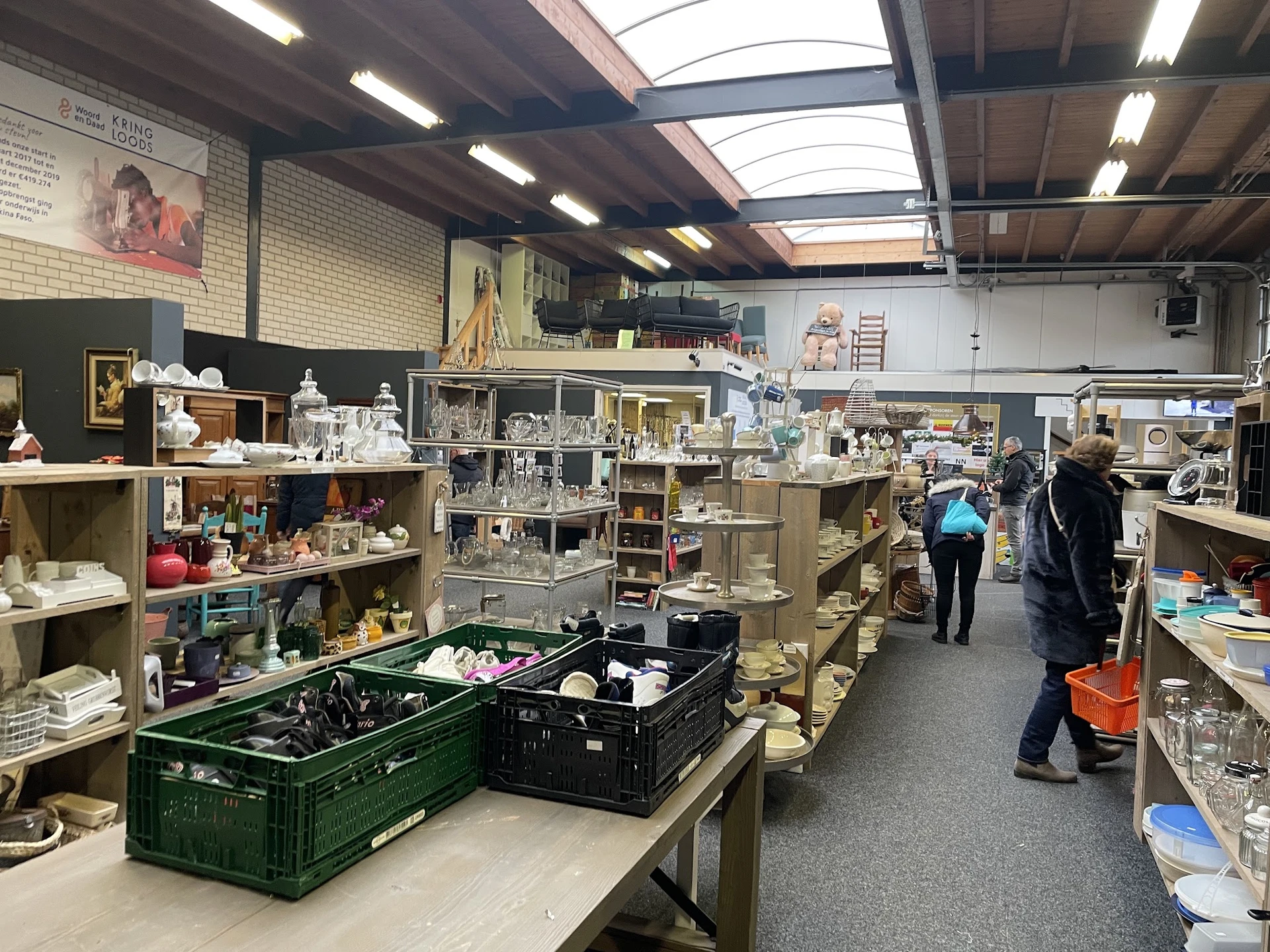 Winkelvloer van kringloopwinkel met stellingen vol spullen, tafels met bakken en bezoekers die rondkijken