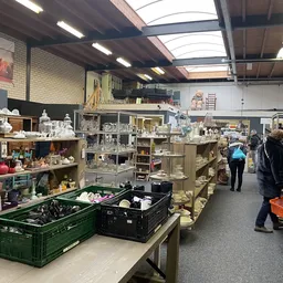 Winkelvloer van kringloopwinkel met stellingen vol spullen, tafels met bakken en bezoekers die rondkijken