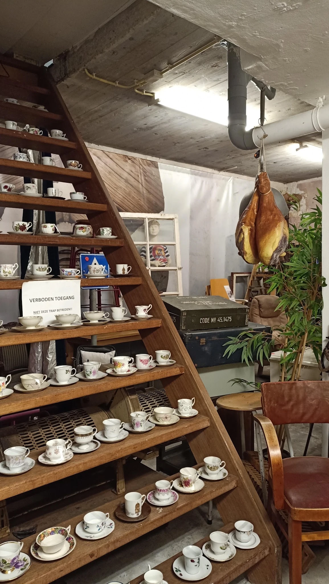 Houten stellingkast met wit servies, kopjes, theepotten en decoratieve items; gedroogde ham hangt ernaast