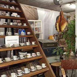 Houten stellingkast met wit servies, kopjes, theepotten en decoratieve items; gedroogde ham hangt ernaast