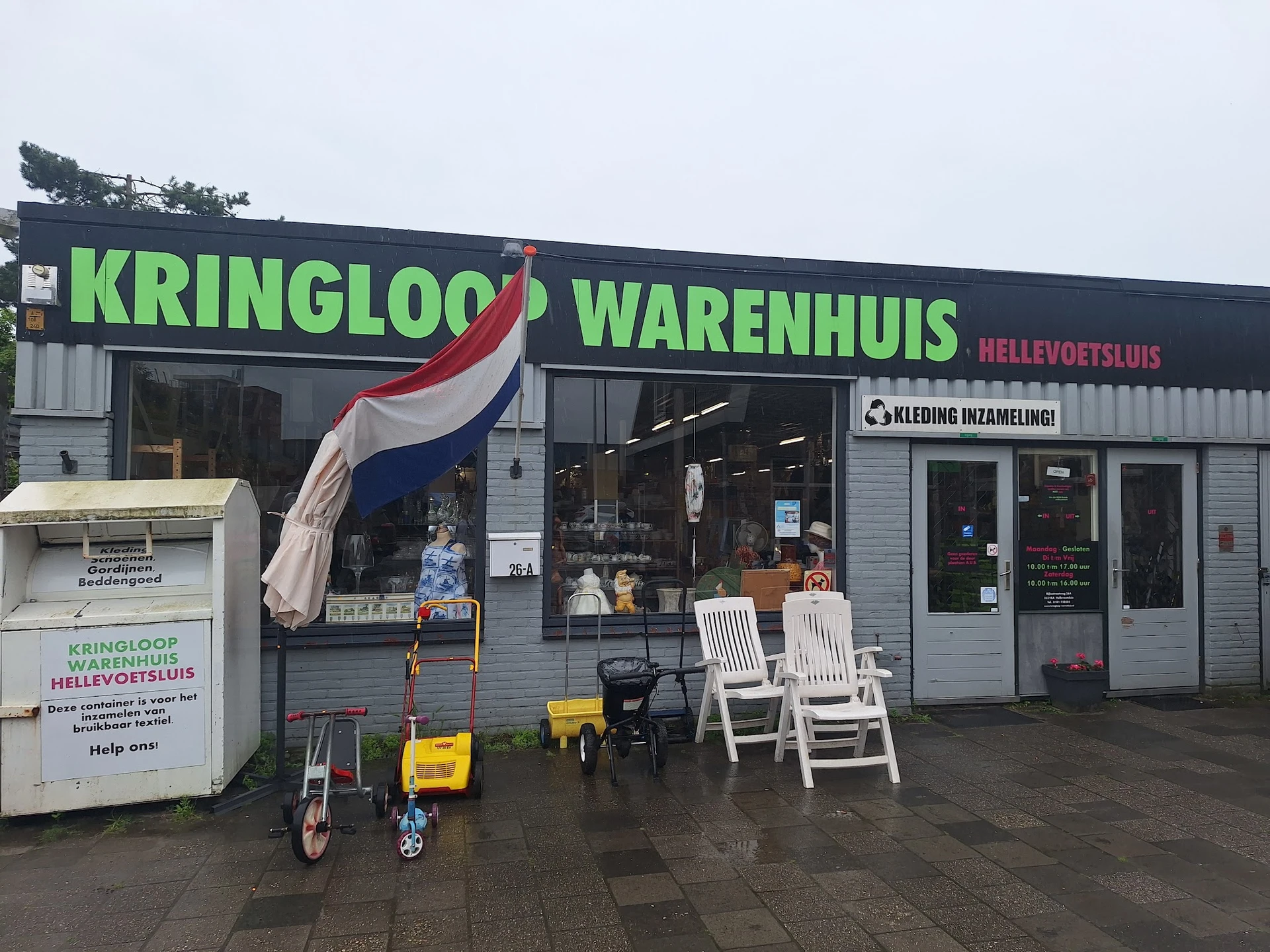 Voorgevel van kringloopwinkel met groene letters, Nederlandse vlag, en witte tuinstoelen op het terras