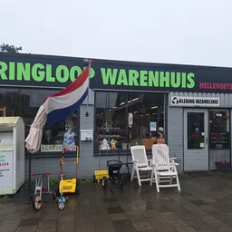 Voorgevel van kringloopwinkel met groene letters, Nederlandse vlag, en witte tuinstoelen op het terras