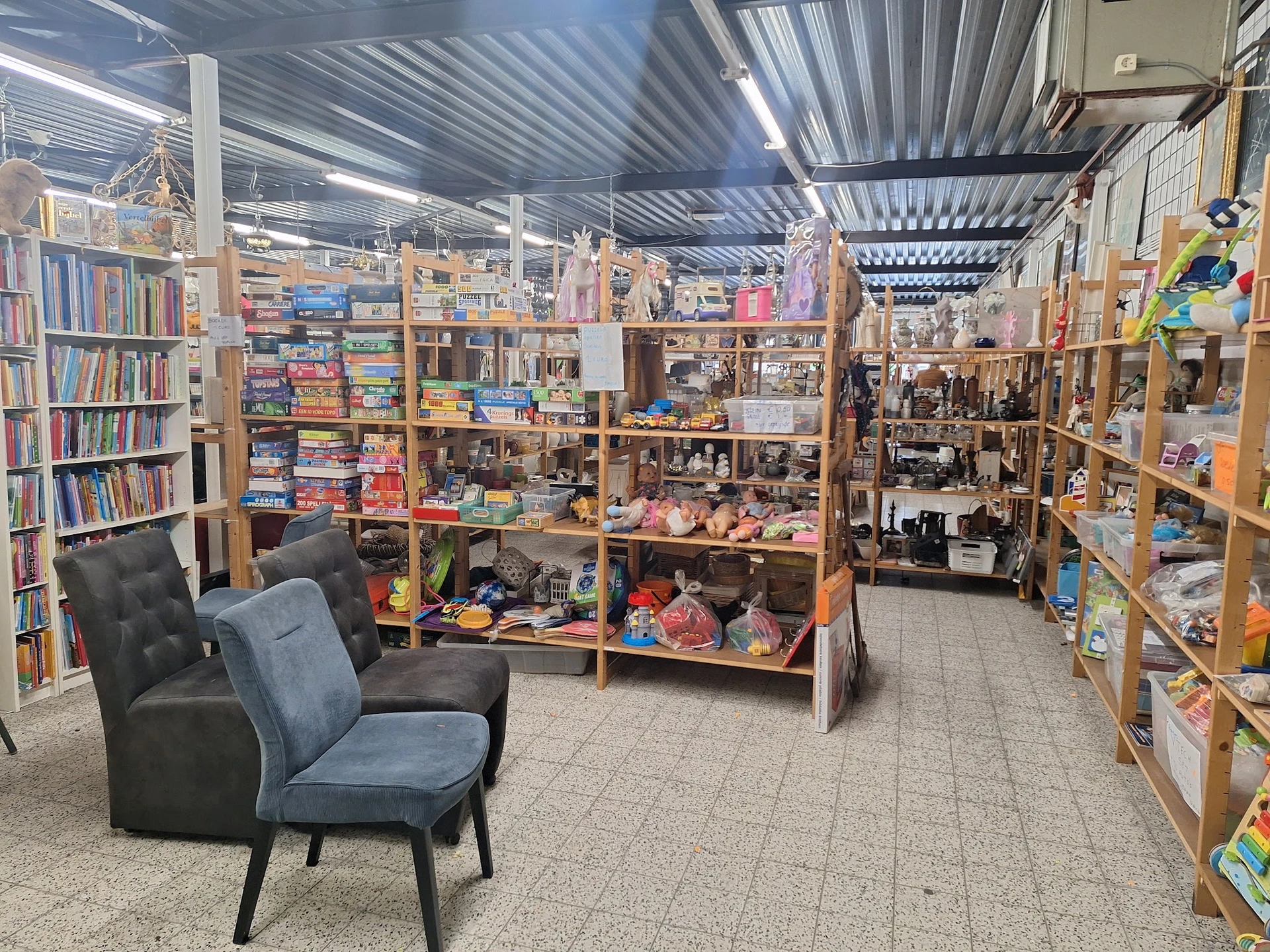 Winkelvloer met houten stellingen vol boeken, speelgoed en diverse spullen, met een zwarte stoel op de voorgrond