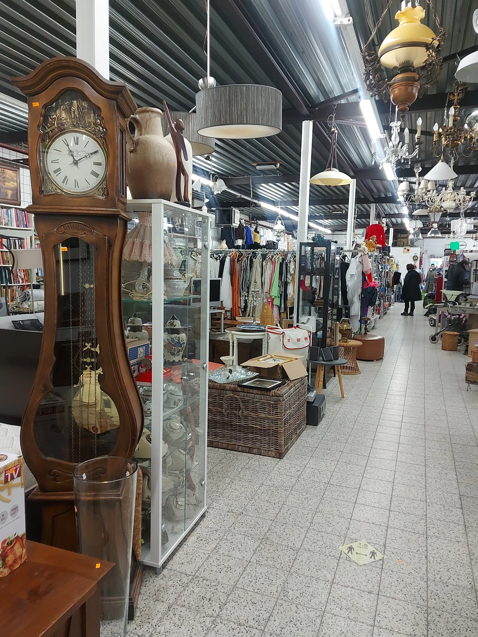 Winkelvloer van kringloopwinkel met stellingen kleding, boeken, meubels en diverse spullen; houten staande klok op voorgrond