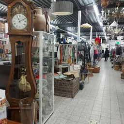 Winkelvloer van kringloopwinkel met stellingen kleding, boeken, meubels en diverse spullen; houten staande klok op voorgrond