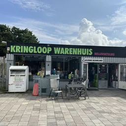 Voorgevel van kringloopwinkel met zwart-wit pui, groene deur en terras met stoelen