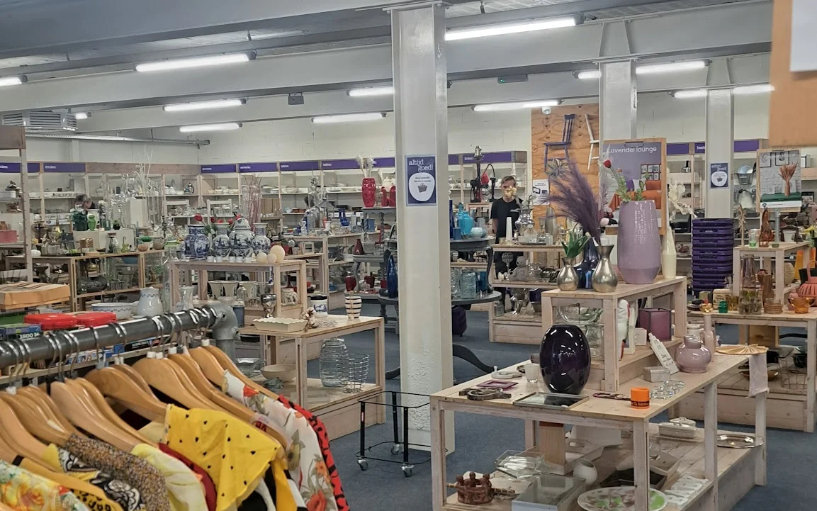 Winkelvloer van kringloopwinkel met kledingrekken, display-tafels met huishoudwaar en decoratie, verlichte plafonds en divers