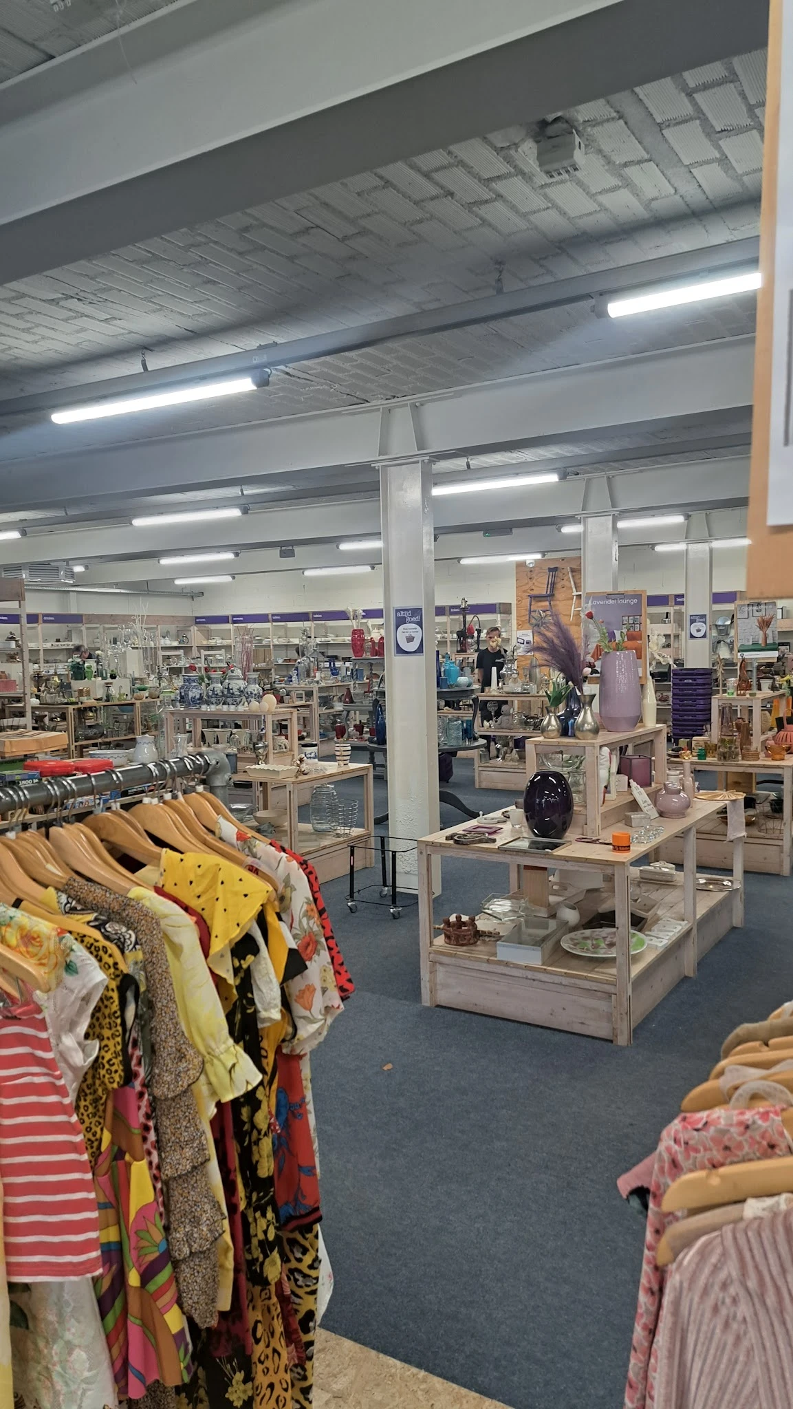 Winkelvloer van kringloopwinkel met kledingrekken, display-tafels met huishoudwaar en decoratie, verlichte plafonds en divers
