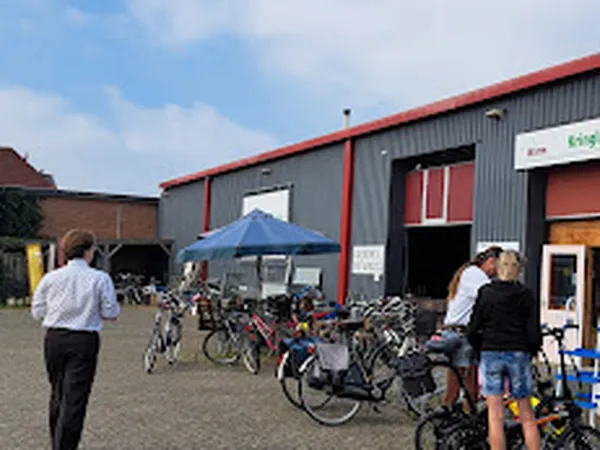 Exterieur van kringloopwinkel met grijze golfplaten gevel, rode accenten en ingang. Voor het pand fietsen geparkeerd en bezoe