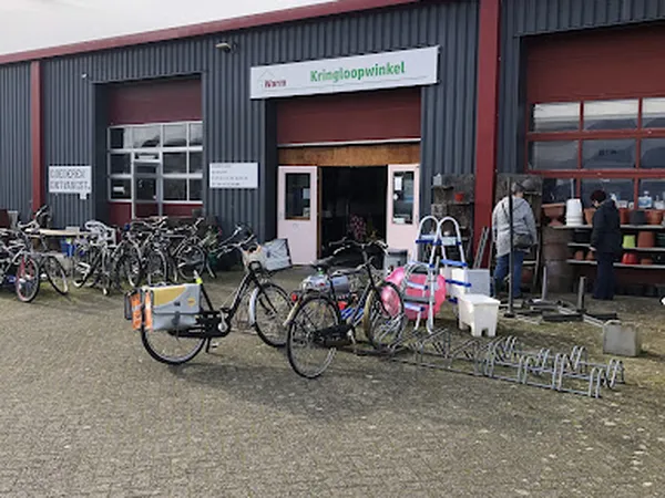Voorkant van kringloopwinkel met ingangsdeur, fietsenstalling en enkele uitgestalde artikelen buiten