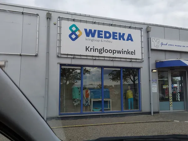 Gevel van kringloopwinkel met WEDEKA-logo, etalage met kleding en blauwe deur