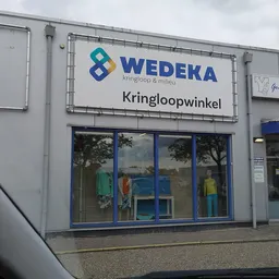 Gevel van kringloopwinkel met WEDEKA-logo, etalage met kleding en blauwe deur