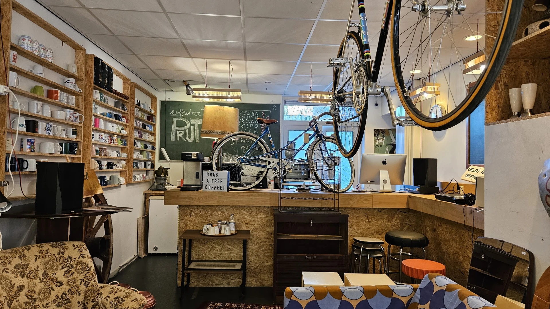 Winkelbinnenruimte met houten toonbank, fietsen aan het plafond, boekenrekken, groene schoolbord en zithoek met oranje access
