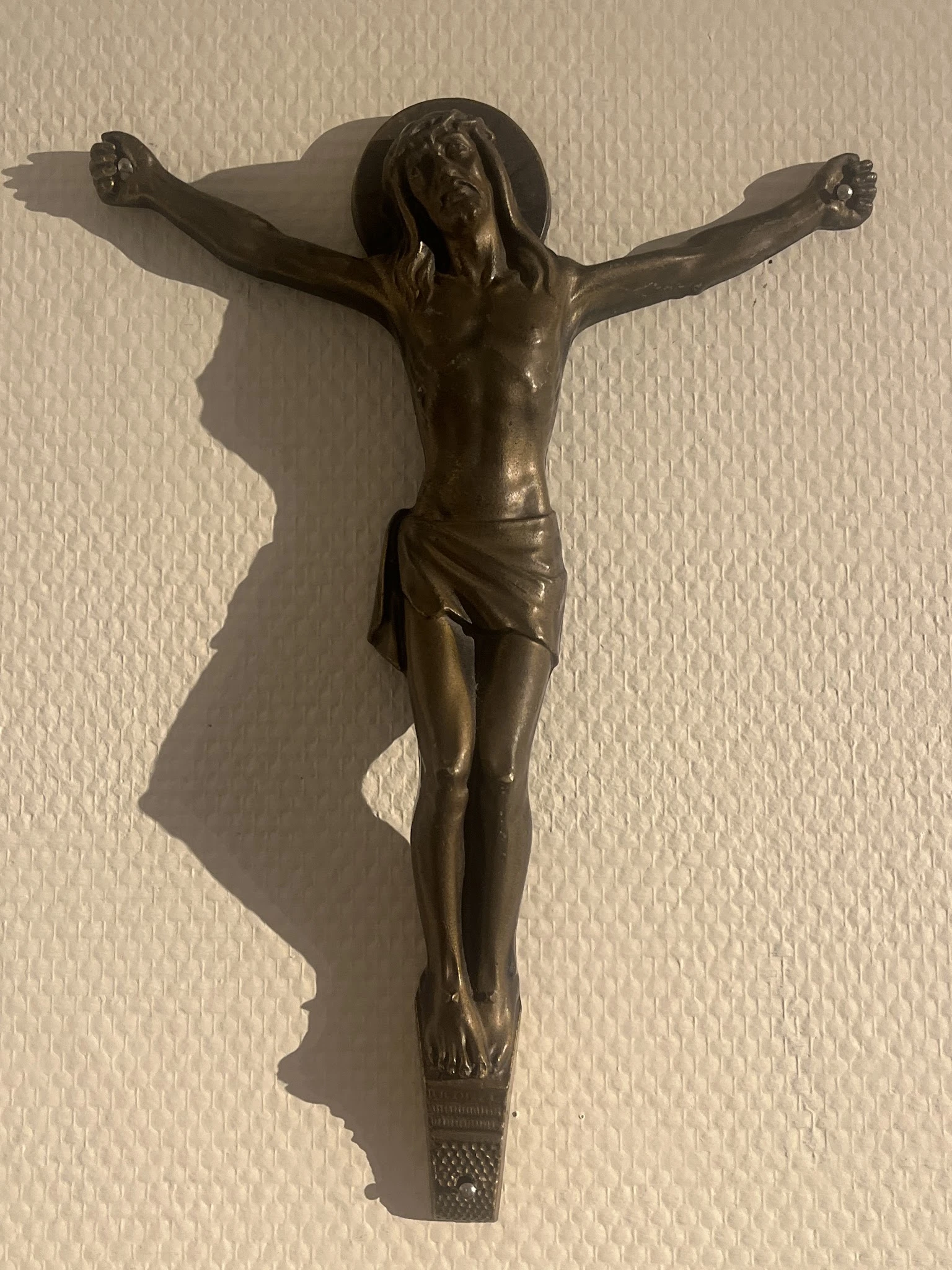 Bronzen crucifix tegen beige muur, met duidelijke schaduwwerking