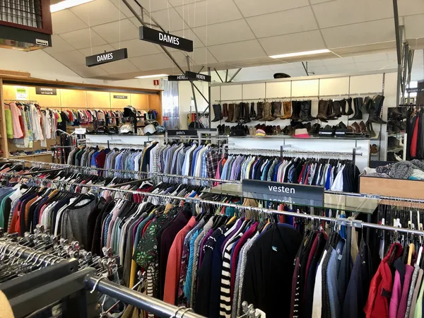 Overzicht van de winkelvloer met rekken vol tweedehands kleding, aangeduid met 'DAMES' bordjes aan het plafond.