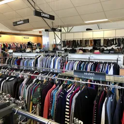 Overzicht van de winkelvloer met rekken vol tweedehands kleding, aangeduid met 'DAMES' bordjes aan het plafond.