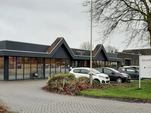 Gevel van kringloopwinkel met grote ramen, zwarte dakkapellen en parkeerplaats met auto's ervoor