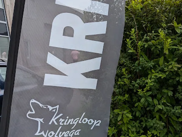 Gevel van kringloopwinkel met witte letters KRINGLOOP WOLVEGA en logo op donkere achtergrond, groen klimop ernaast