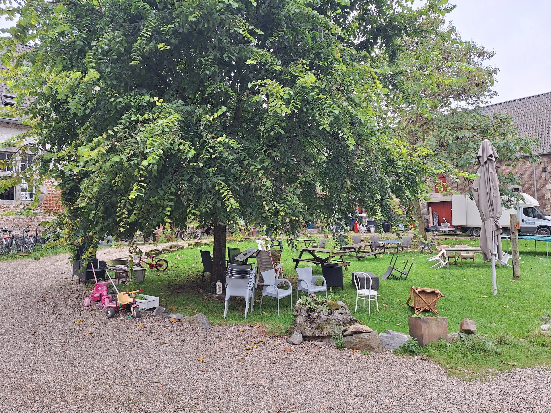 Terras met tafels en stoelen onder grote boom in groene tuin, met gebouwen op achtergrond