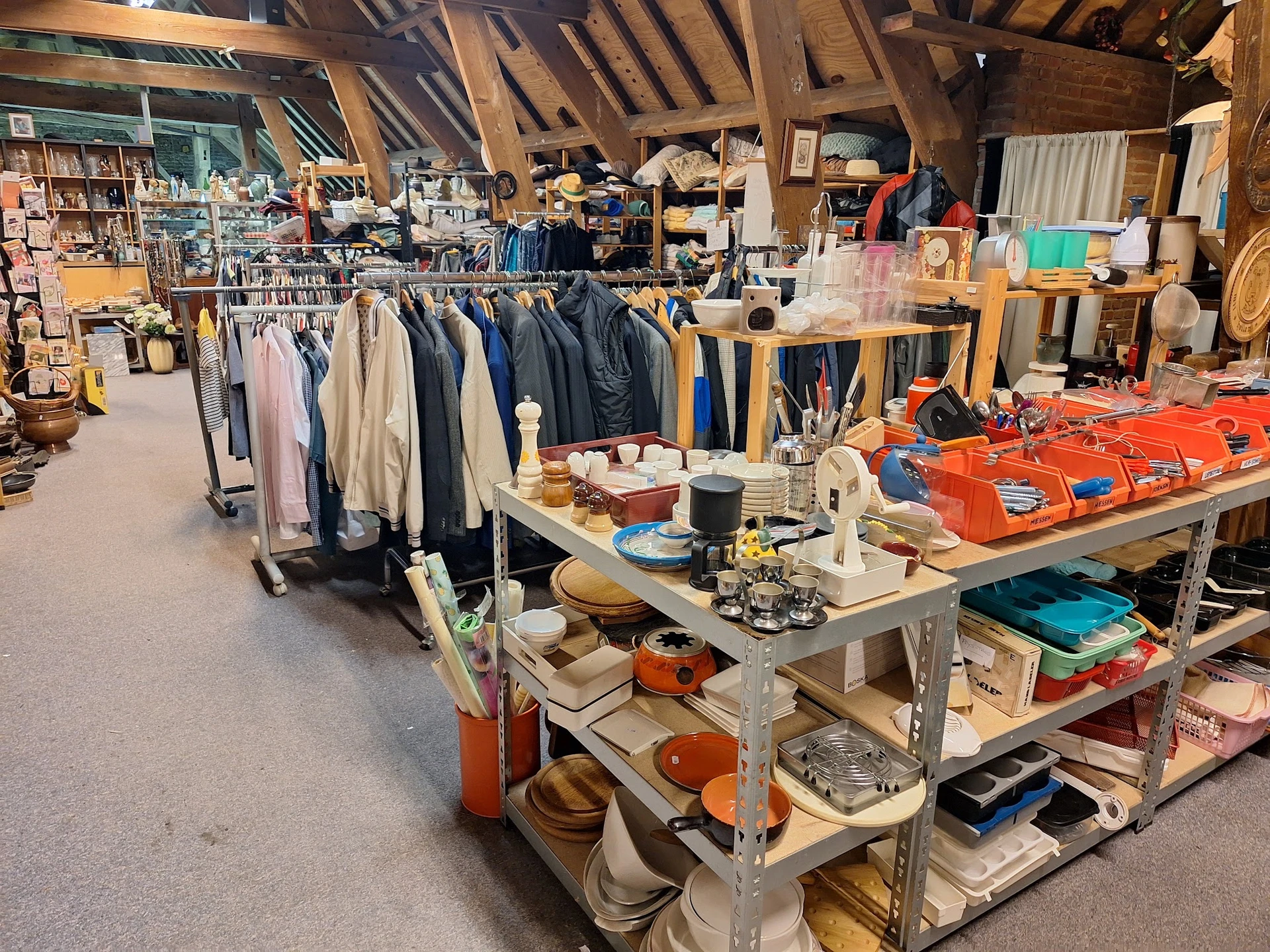 Overzicht van kringloopwinkel met kledingrekken, werktafels en diverse spullen op zolderachtige ruimte
