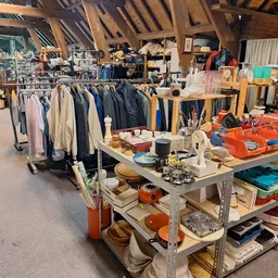 Overzicht van kringloopwinkel met kledingrekken, werktafels en diverse spullen op zolderachtige ruimte