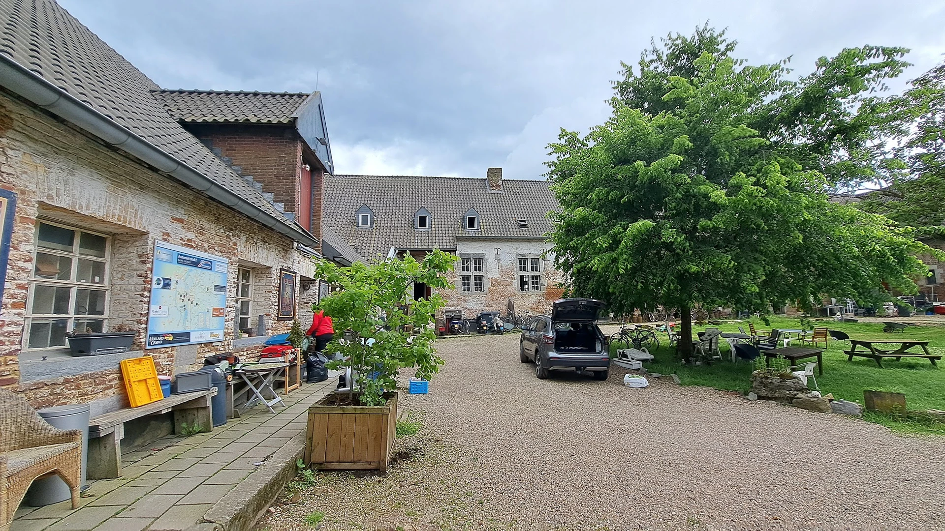 Voorgevel van stenen kringloopwinkelpand met ingang links, grind parkeerterrein en groene bomen