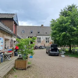 Voorgevel van stenen kringloopwinkelpand met ingang links, grind parkeerterrein en groene bomen