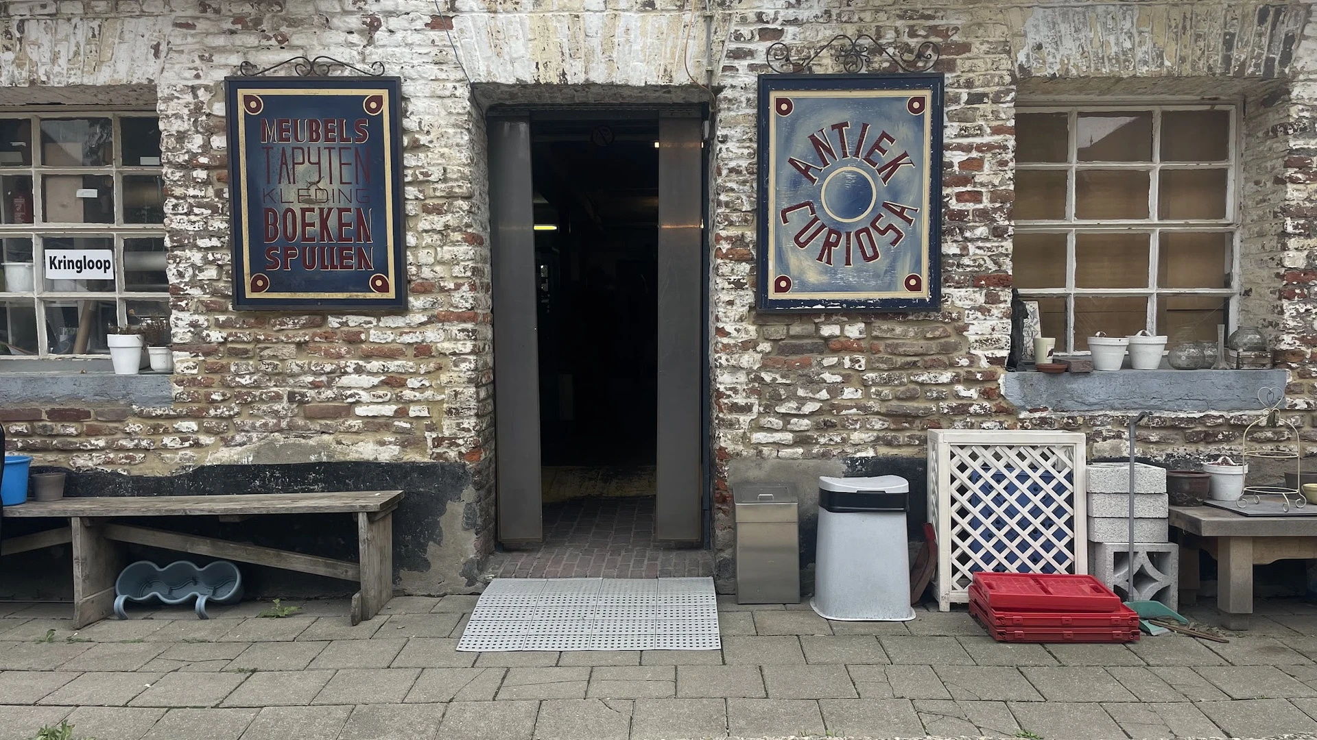 Ingang van kringloopwinkel met twee borden aan bakstenen gevel, bank links en diverse items op straat