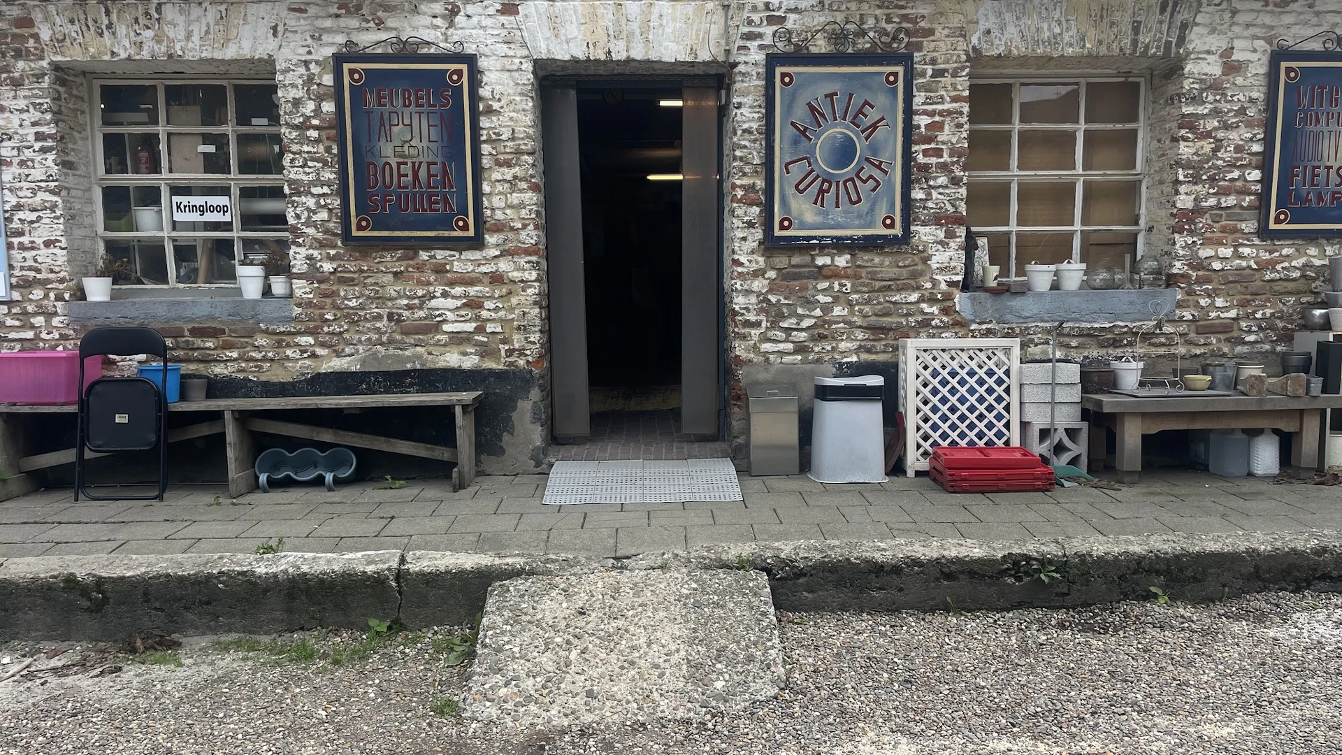 Voorgevel van kringloopwinkel met bakstenen muur, zwarte deur, twee blauwe borden en diverse spullen op de stoep