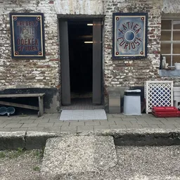 Voorgevel van kringloopwinkel met bakstenen muur, zwarte deur, twee blauwe borden en diverse spullen op de stoep