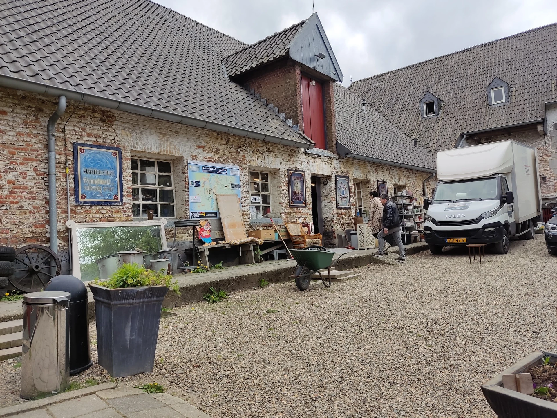 Bakstenen gevel van kringloopwinkel met ingang, uitstallingen en parkeerplaats met witte bestelbus
