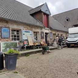 Bakstenen gevel van kringloopwinkel met ingang, uitstallingen en parkeerplaats met witte bestelbus