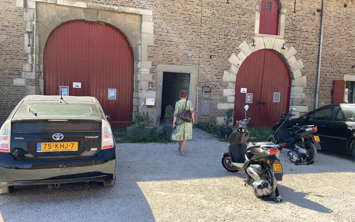 Bakstenen gevel van kringloopwinkel met twee rode boogdeuren, parkeerplaats met auto's en scooters