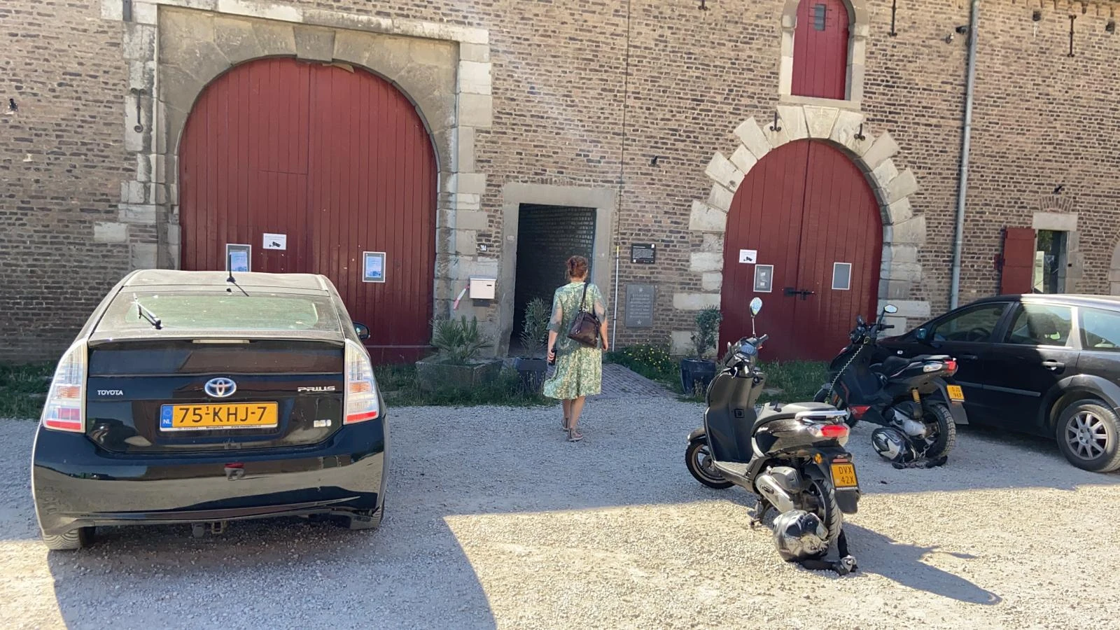 Bakstenen gevel van kringloopwinkel met twee rode boogdeuren, parkeerplaats met auto's en scooters