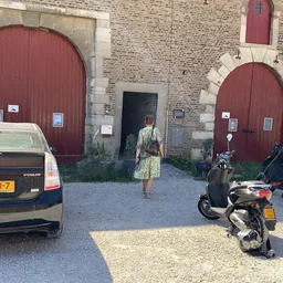 Bakstenen gevel van kringloopwinkel met twee rode boogdeuren, parkeerplaats met auto's en scooters