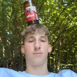 Portretfoto van een jongen met een rode beker op zijn hoofd, tegen groene bladeren