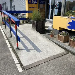 Toegangspad met blauwe reling naar winkelingang met gele en blauwe elementen
