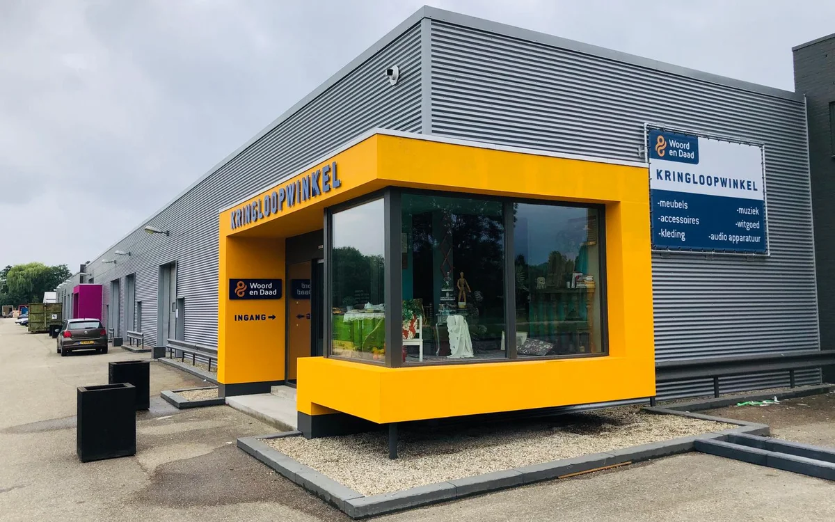 Gele ingang van kringloopwinkel aan grijze loods met grote ramen en signage