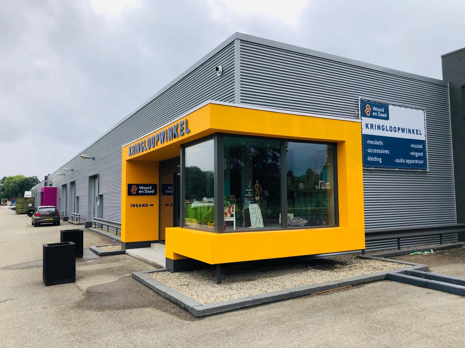 Gele ingang van kringloopwinkel aan grijze loods met grote ramen en signage