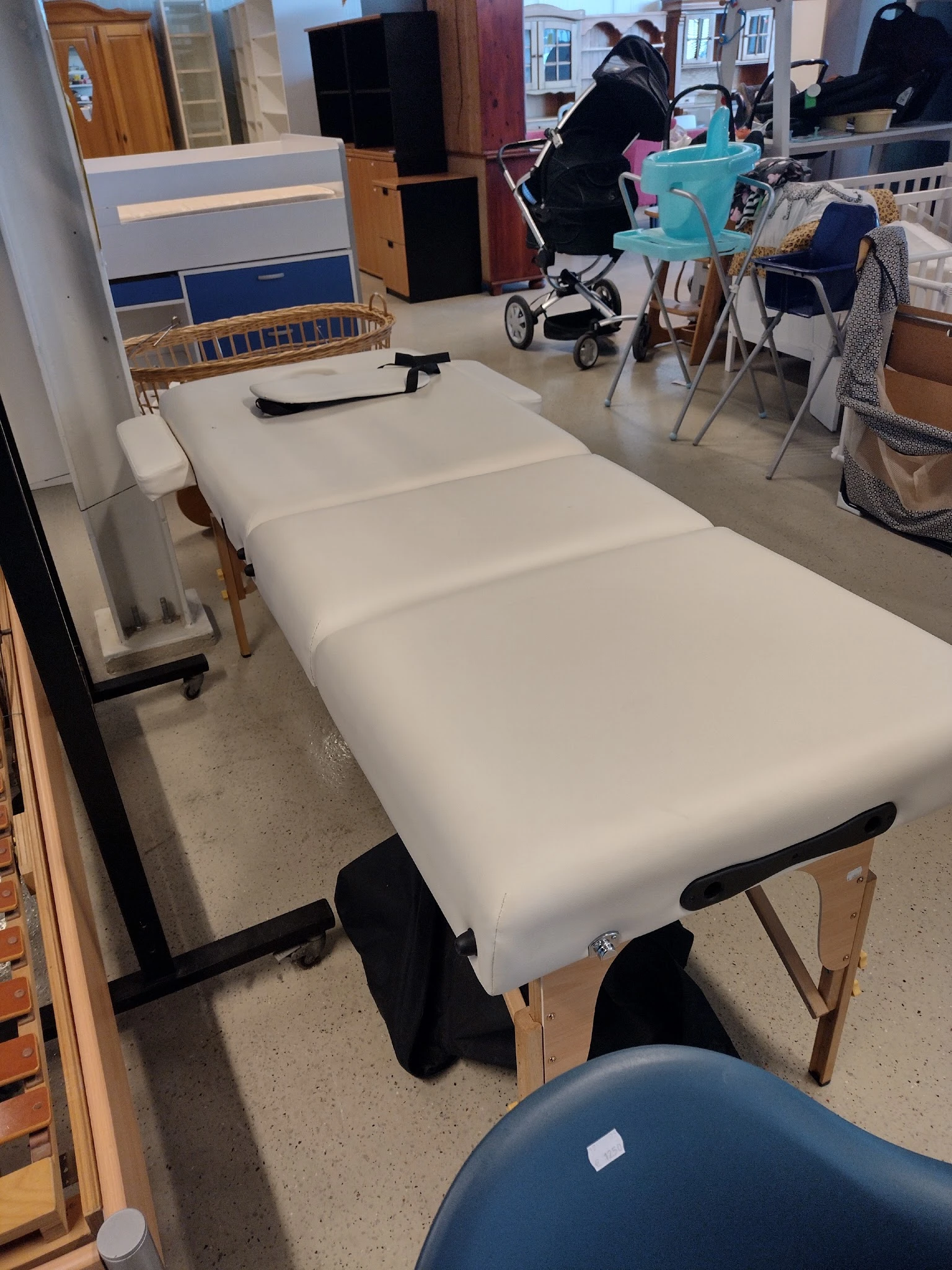 Wit massagetafel of behandeltafel met zwarte poten, opgesteld in een winkelruimte met speelgoed en babyspeelgoed op de achter