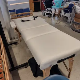 Wit massagetafel of behandeltafel met zwarte poten, opgesteld in een winkelruimte met speelgoed en babyspeelgoed op de achter