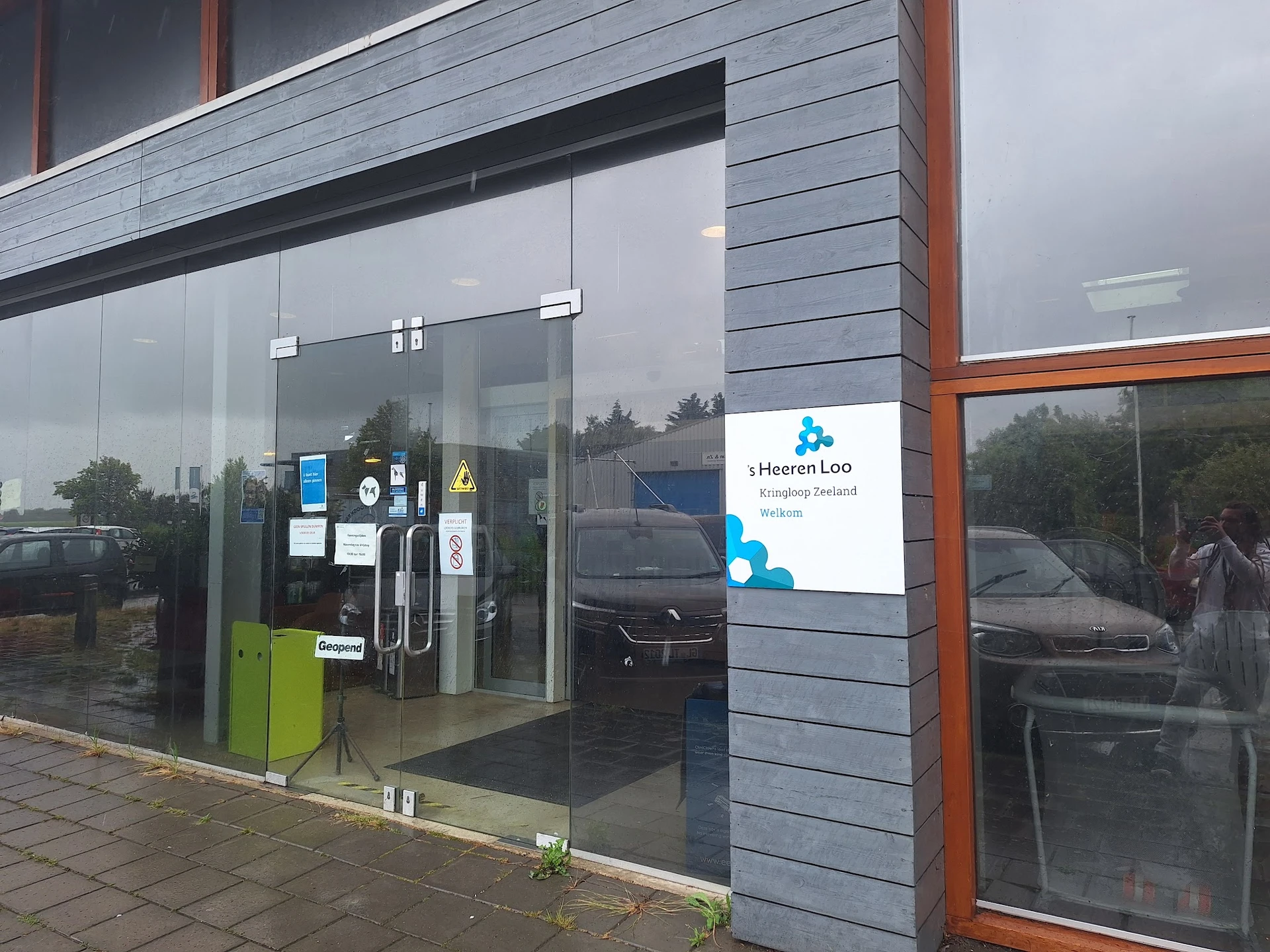 Moderne winkelpui met glazen deuren en ramen, met blauw logo en witte zijgevel met bruine palen