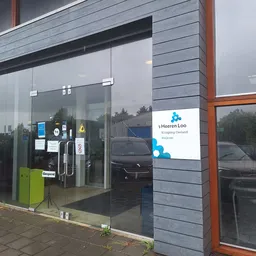 Moderne winkelpui met glazen deuren en ramen, met blauw logo en witte zijgevel met bruine palen
