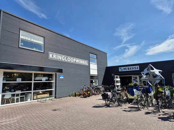 Gevel van kringloopwinkel met naam KRINGLOOPWINKEL in letters op grijze gevelbekleding, vitrine met spullen en fietsenstallin