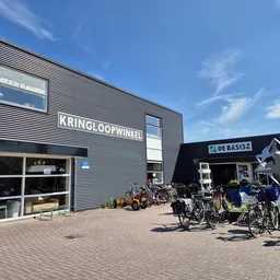 Gevel van kringloopwinkel met naam KRINGLOOPWINKEL in letters op grijze gevelbekleding, vitrine met spullen en fietsenstallin