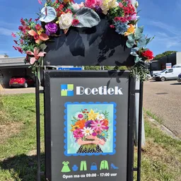 Zwarte brievenbus met Boetiek-logo en bloemen ernaast buiten de winkel
