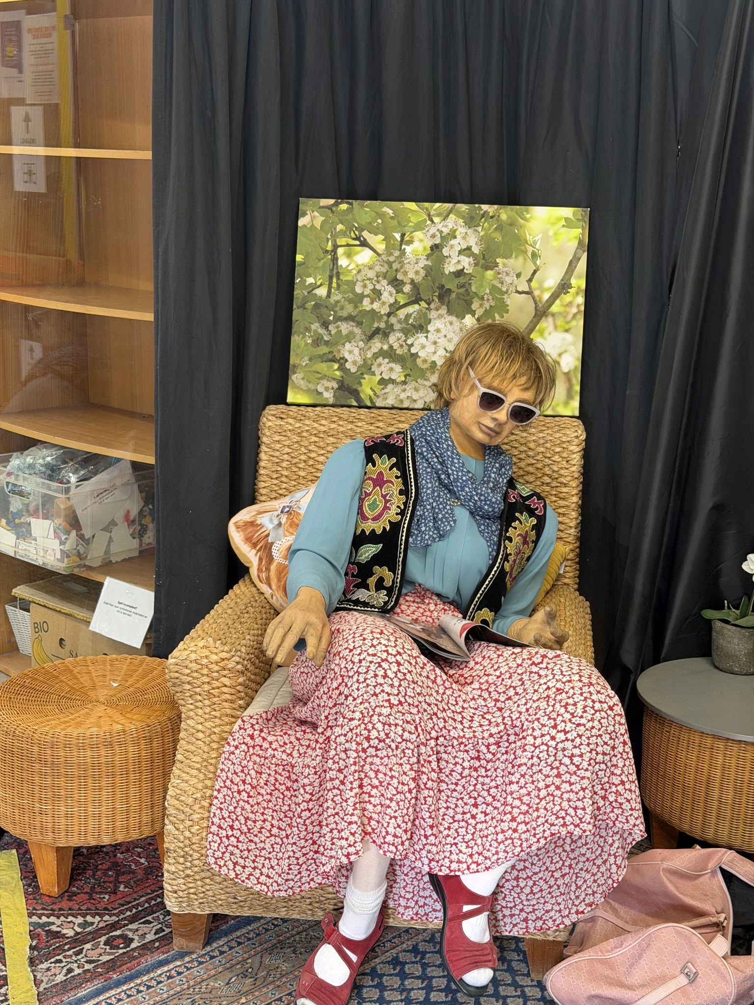 Mannequin in vintage outfit zittend op rieten stoel in winkelhoek met zwarte gordijnen en houten stellingen op achtergrond