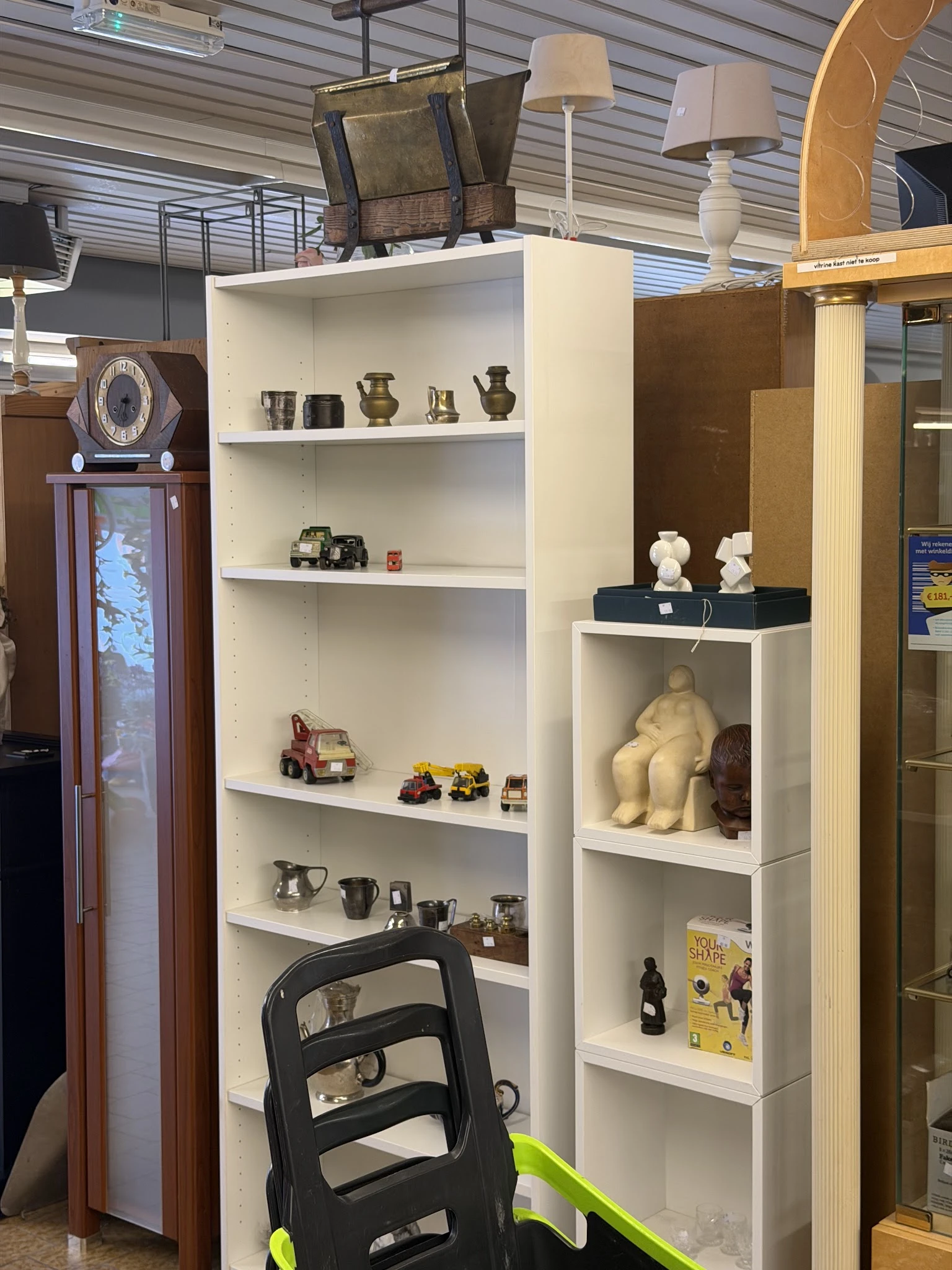 Witte wandkasten met diverse tweedehands items: metalen voorwerpen, speelgoed, beelden en decoratieve spullen.