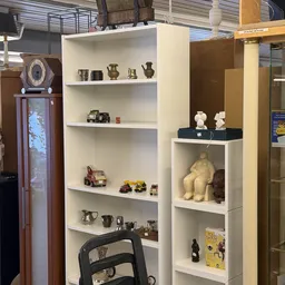 Witte wandkasten met diverse tweedehands items: metalen voorwerpen, speelgoed, beelden en decoratieve spullen.